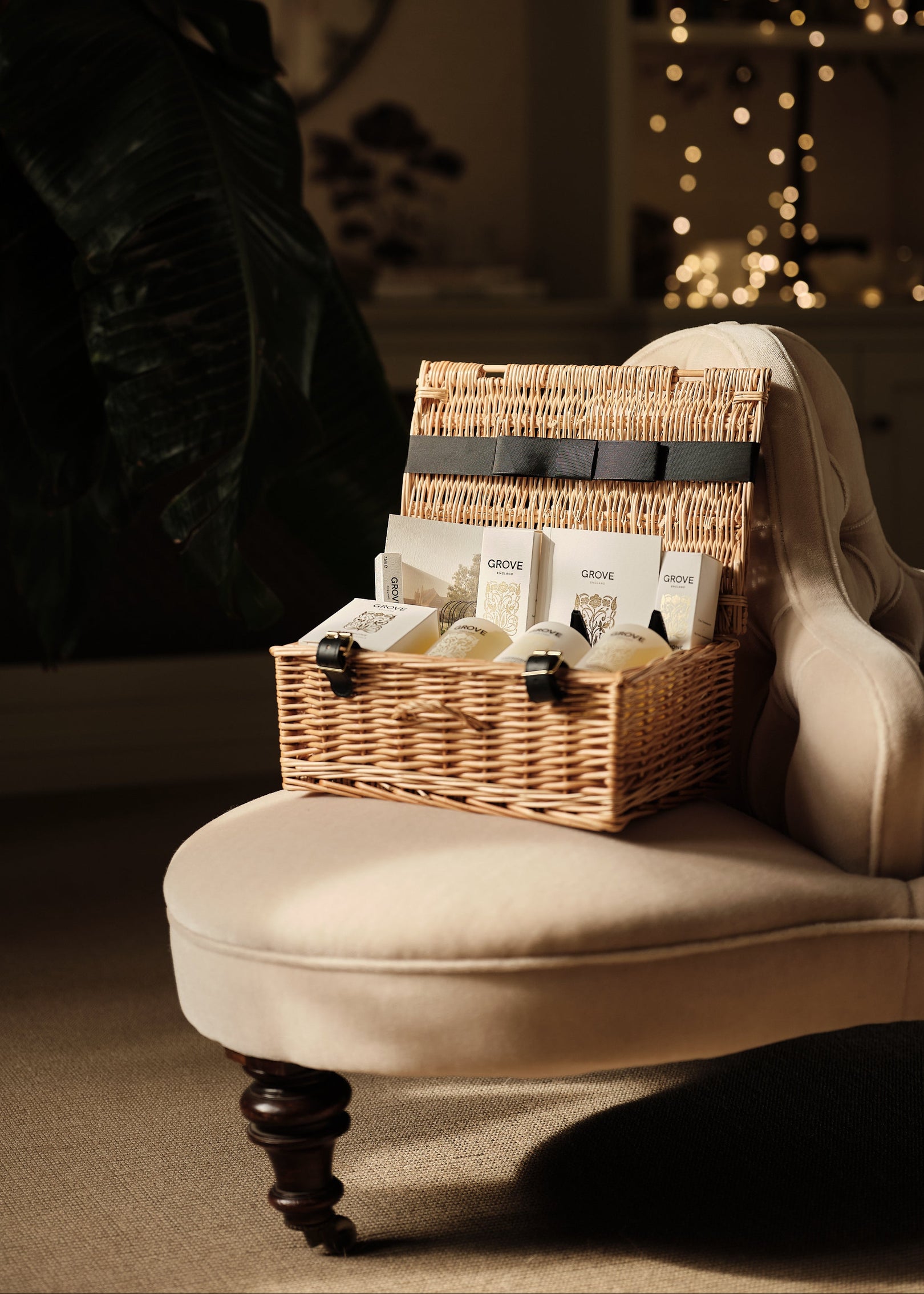 Wicker gift basket with items on a beige sofa in a dimly lit room with plants and string lights.