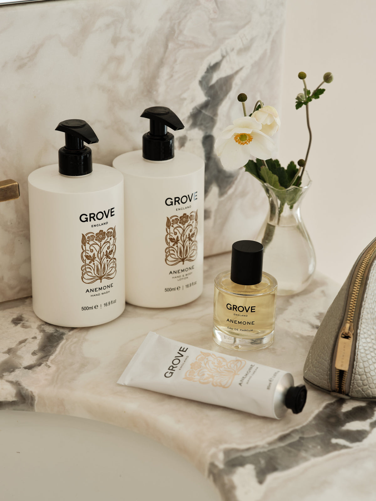 A GROVE Anemone body care set arranged on a marble surface, including two large white pump bottles, a perfume bottle, a white hand cream tube, and a textured light grey pouch, styled with a small vase of white flowers.