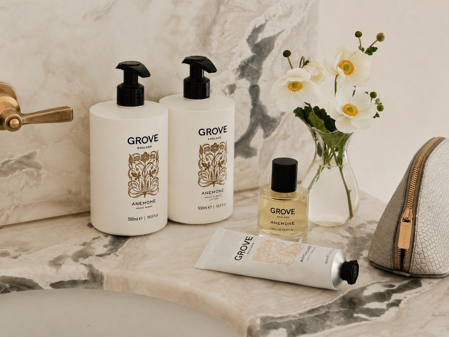 Anemone by GROVE England bathroom and body care set arranged on a marble sink, including two large pump bottles, a small perfume bottle, a hand cream tube, and a textured zip pouch, with white flowers in a glass vase nearby.
