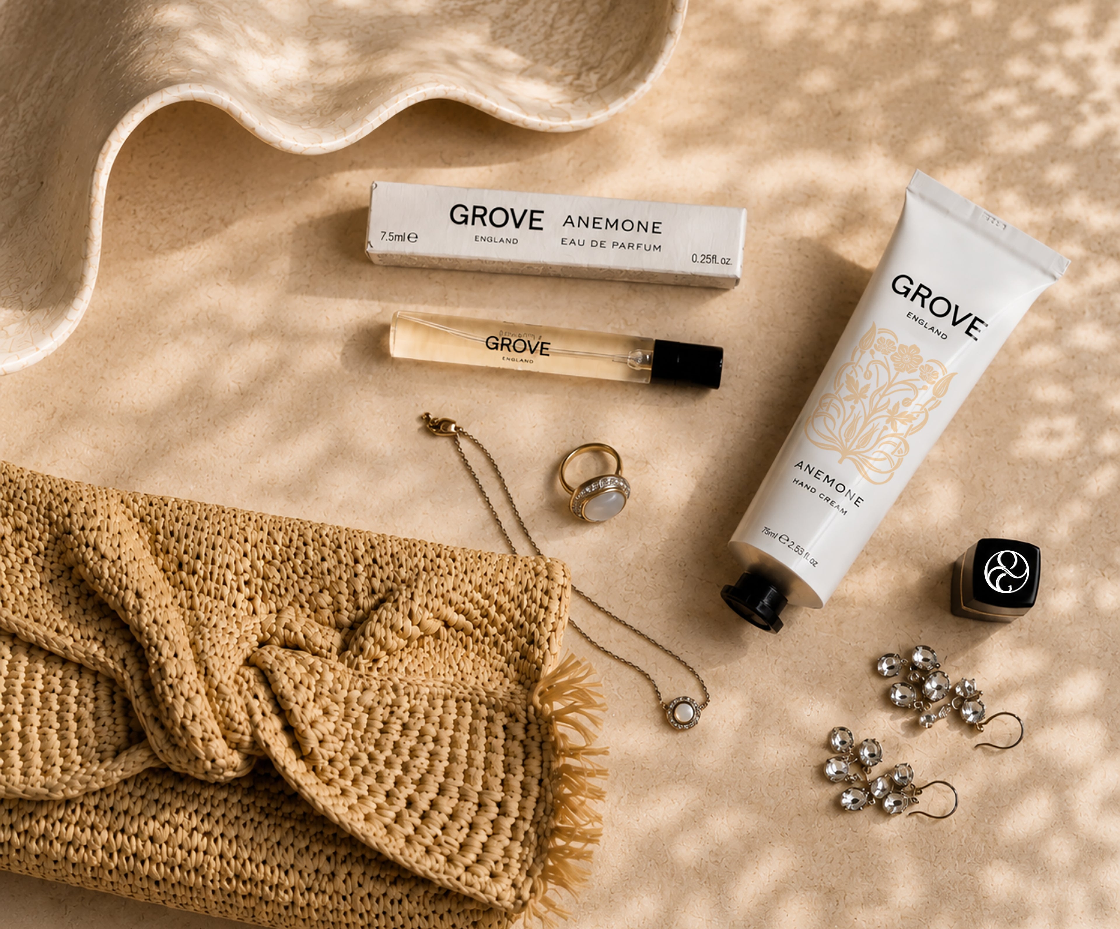 A flat-lay arrangement of GROVE Anemone perfume and fragrance products on a sandy textured surface with dappled light. Visible items include a white box, a small glass perfume vial, a white hand cream tube, styled with a woven clutch, and jewelry