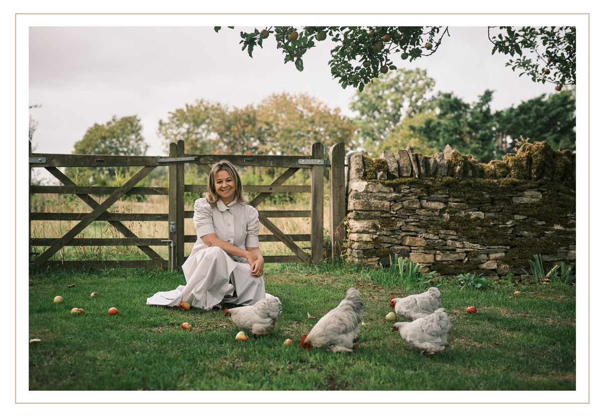 Laura sits on the grassy lawn  of the orchard in front of a wooden fence and stone wall, with several Lavender Pekin chickens pecking at the ground around her.