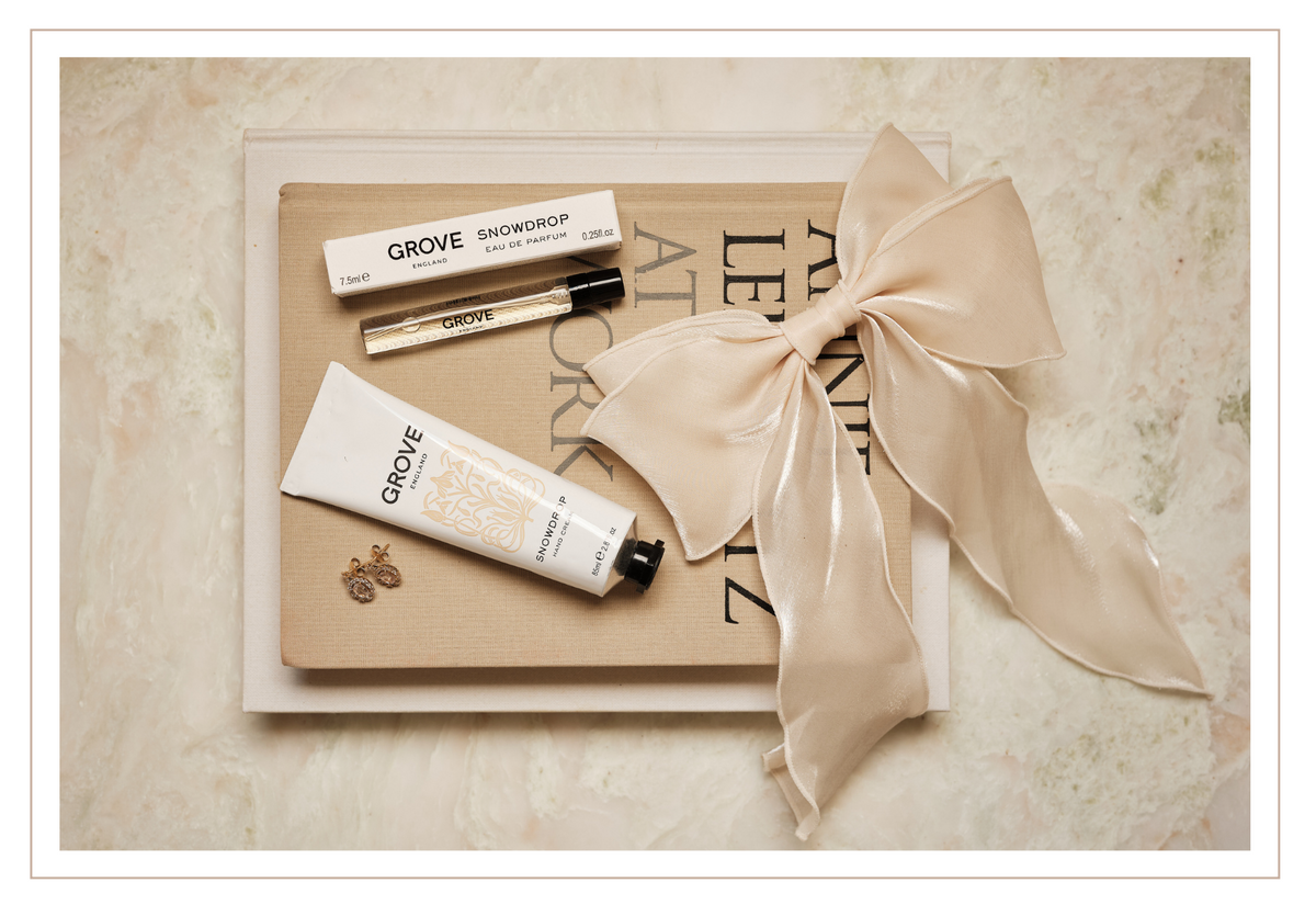 GROVE England Scent Duo gift set arranged on a beige book, featuring a Snowdrop Hand Cream, a small vial of Snowdrop Eau de Parfum, and a large cream satin ribbon bow, photographed from above on a light marbled surface.