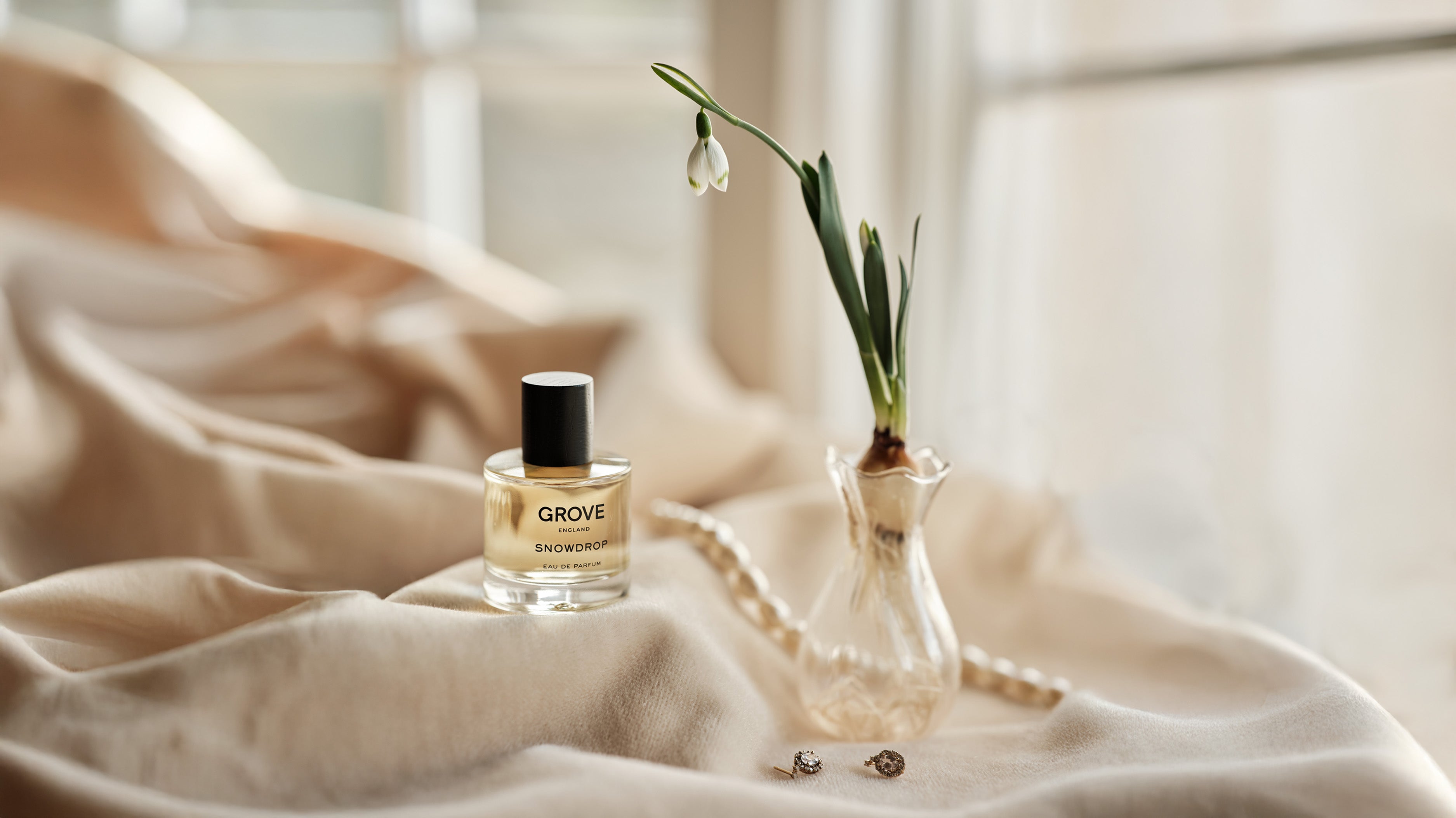 Perfume bottle labeled 'Grove' on a soft velvet surface with a small bud vase holding a Snowdrop, and pearls in the background.
