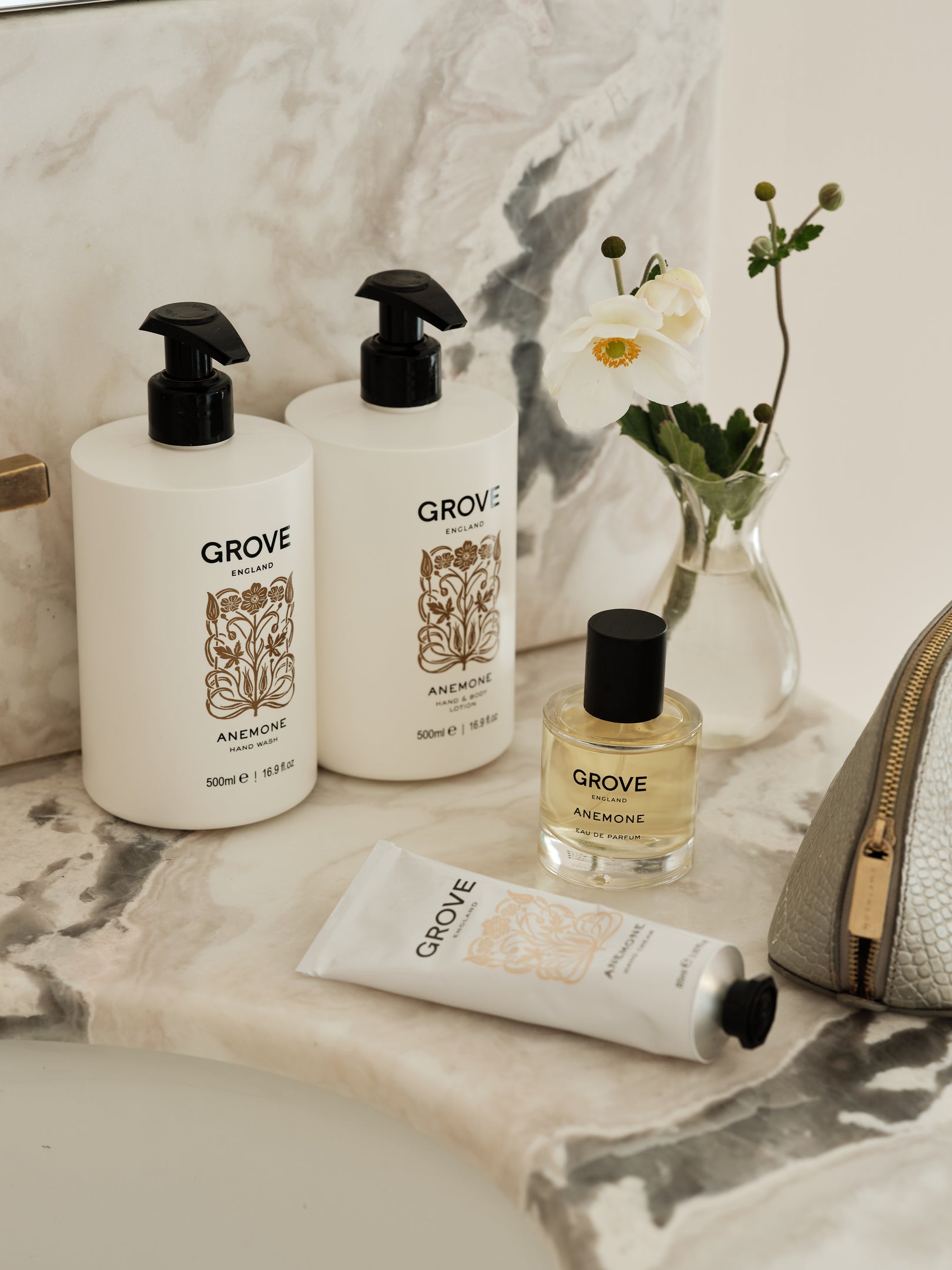 A GROVE Anemone body care set arranged on a marble surface, including two large white pump bottles, a perfume bottle, a white hand cream tube, and a textured light grey pouch, styled with a small vase of white flowers.